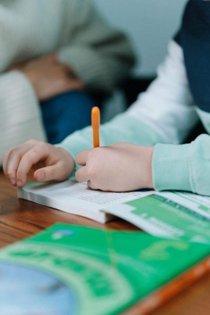 Close-up of a child's hands writing with an orange pen on a book indoors.